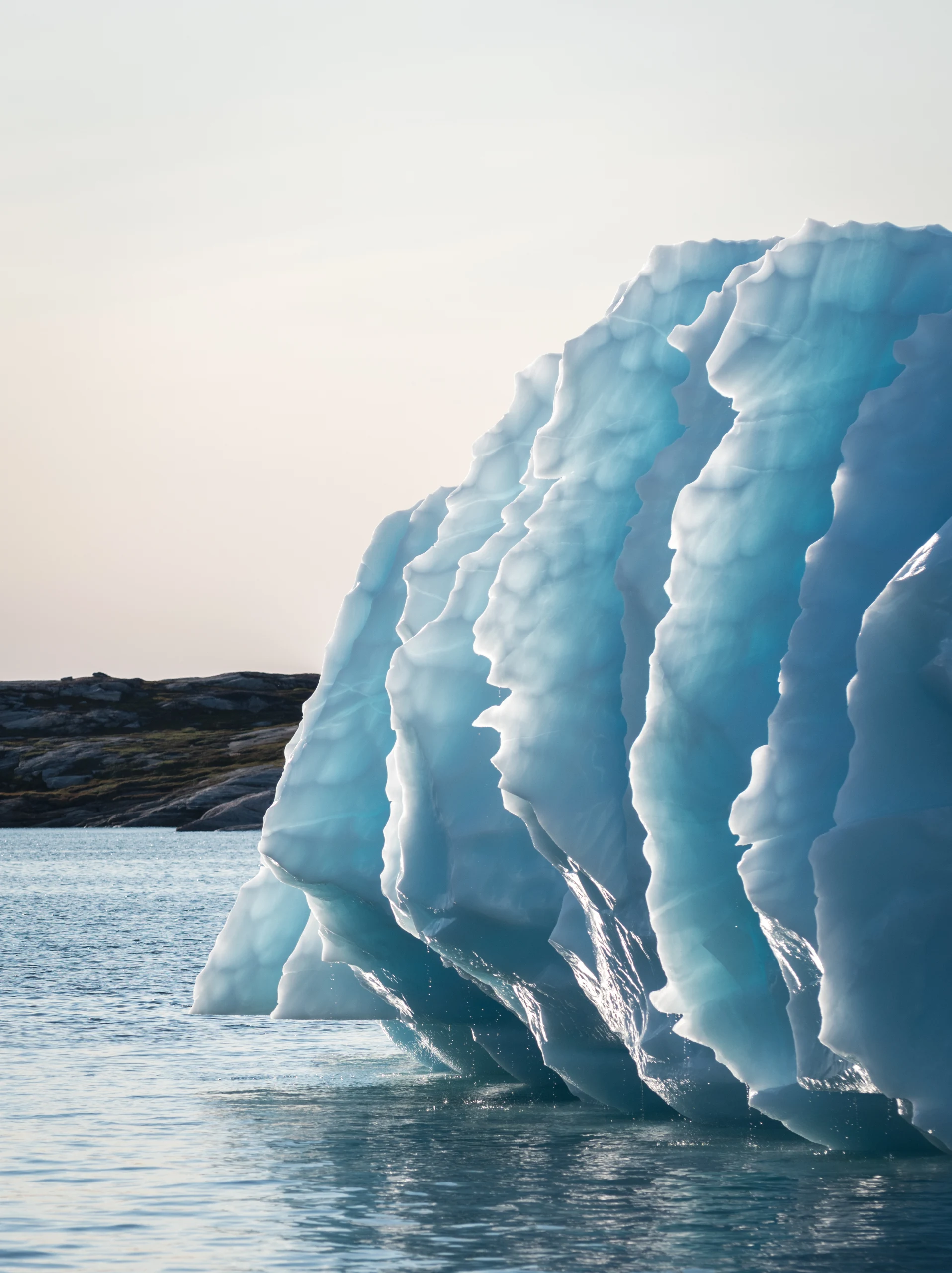A blue iceberg face, deeply scalloped by summer melt, rises above calm water in Greenland.